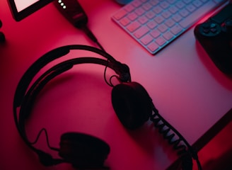 A dimly lit gaming setup with a monitor displaying a game's menu on the screen. A mechanical keyboard sits in front of the monitor. A pair of over-ear headphones and a game controller rest on the desk. The scene is bathed in a red and blue light, creating a moody gaming atmosphere.