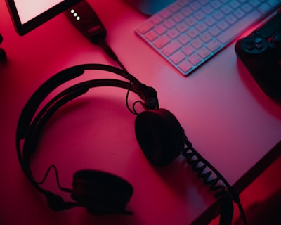A dimly lit gaming setup with a monitor displaying a game's menu on the screen. A mechanical keyboard sits in front of the monitor. A pair of over-ear headphones and a game controller rest on the desk. The scene is bathed in a red and blue light, creating a moody gaming atmosphere.