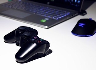 A black gaming console controller is placed on a light surface next to a sleek laptop and a gaming mouse with a blue LED light. The setting suggests a gaming or tech workspace with a focus on modern electronic devices.