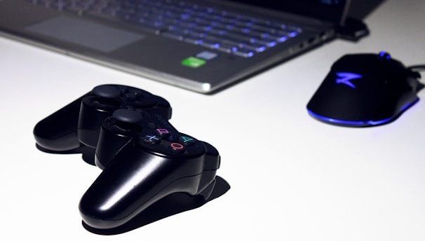 A black gaming console controller is placed on a light surface next to a sleek laptop and a gaming mouse with a blue LED light. The setting suggests a gaming or tech workspace with a focus on modern electronic devices.