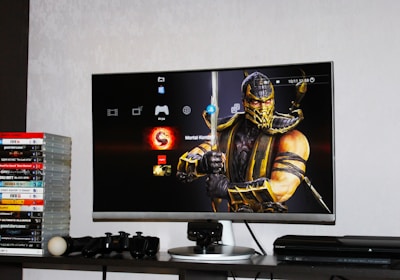 A flat-screen TV displays a video game interface featuring a character from Mortal Kombat in the center. Beside the TV is a stack of video game cases for the PlayStation 3, including titles like FIFA 14 and Grand Theft Auto. Below the TV, there is a gaming console, controllers, and other gaming equipment.
