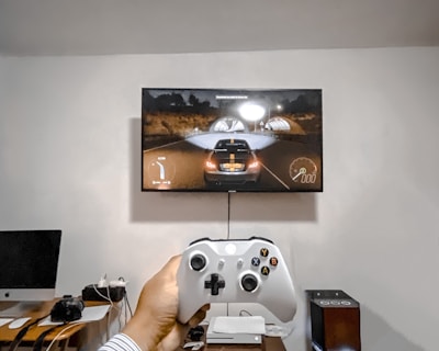 A person is holding a gaming controller in front of a flat-screen television displaying a racing video game. The room has a computer setup on a wooden desk with cables, a desktop computer monitor, and other accessories. The focus is on the interaction between the person and the gaming environment.
