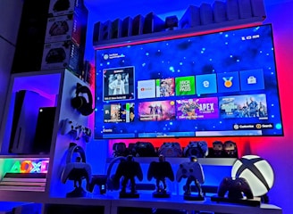 A modern gaming setup featuring a large wall-mounted screen displaying a gaming console interface. The room is decorated with gaming-themed items such as controllers and collectibles. The ambient lighting includes shades of blue and red, creating a vibrant atmosphere.