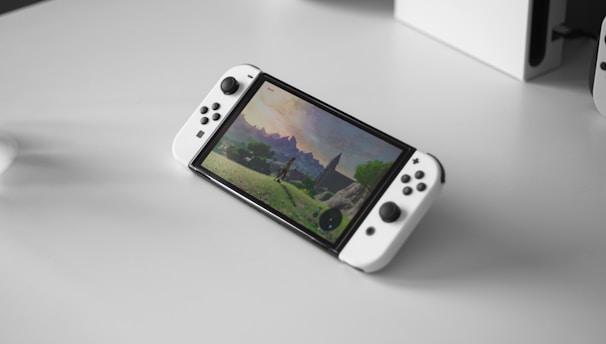 A portable gaming console with a screen showing a video game landscape, resting on a light-colored surface. The console has white controllers attached to either side of the screen. Nearby, there seems to be a white docking station or accessory.