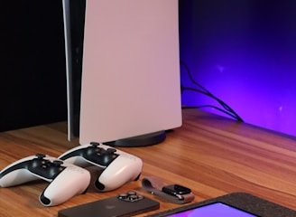 A sleek, modern setup featuring a gaming console with two controllers on a wooden surface. Nearby are a smartphone, smartwatch, tablet with a vivid screen, wireless earbuds, and a stylus. The background is illuminated with a soft purple glow.