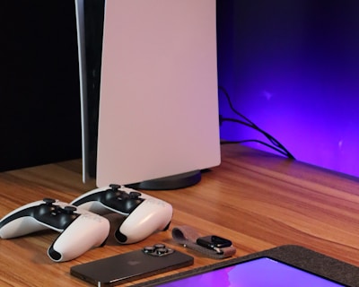 A sleek, modern setup featuring a gaming console with two controllers on a wooden surface. Nearby are a smartphone, smartwatch, tablet with a vivid screen, wireless earbuds, and a stylus. The background is illuminated with a soft purple glow.