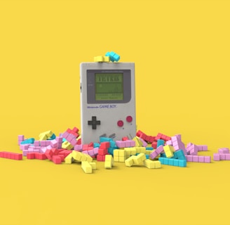 A vintage handheld gaming console surrounded by colorful, block-like pieces reminiscent of a classic puzzle game. The console has a small screen displaying a game in progress, with two red buttons and a directional pad. The vibrant pieces are scattered around it, creating a playful and nostalgic atmosphere.