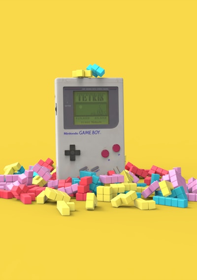 A vintage handheld gaming console surrounded by colorful, block-like pieces reminiscent of a classic puzzle game. The console has a small screen displaying a game in progress, with two red buttons and a directional pad. The vibrant pieces are scattered around it, creating a playful and nostalgic atmosphere.